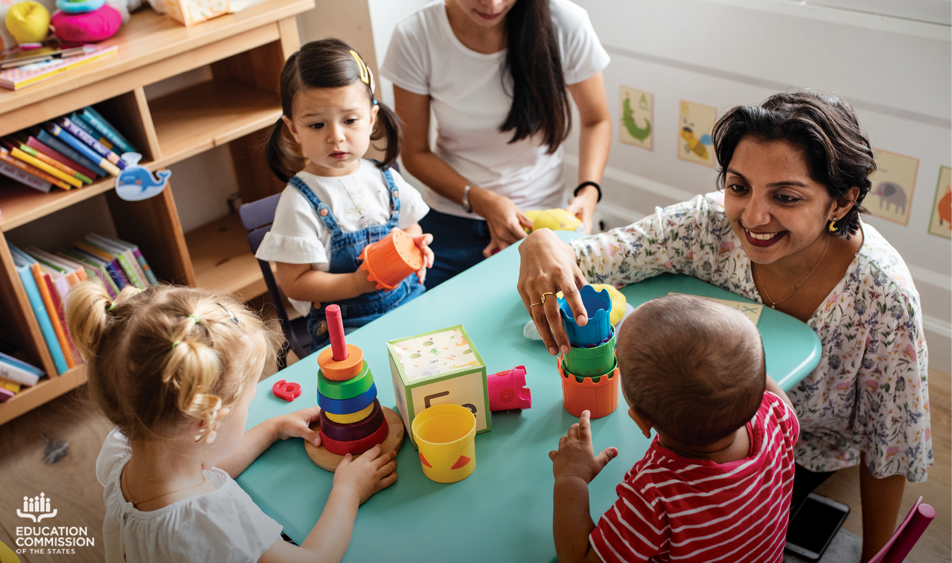 Three young children play with blocks and other toys at a small blue table while two adults assist them. The adult in the frame (who presents as a woman) has short, dark brown hair, olive skin and is wearing a white shirt with colorful flower designs.