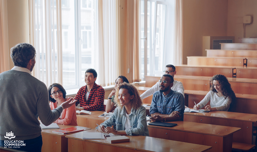 A professor lectures to seven undergraduate students of diverse racial backgrounds in a lecture hall during the day.