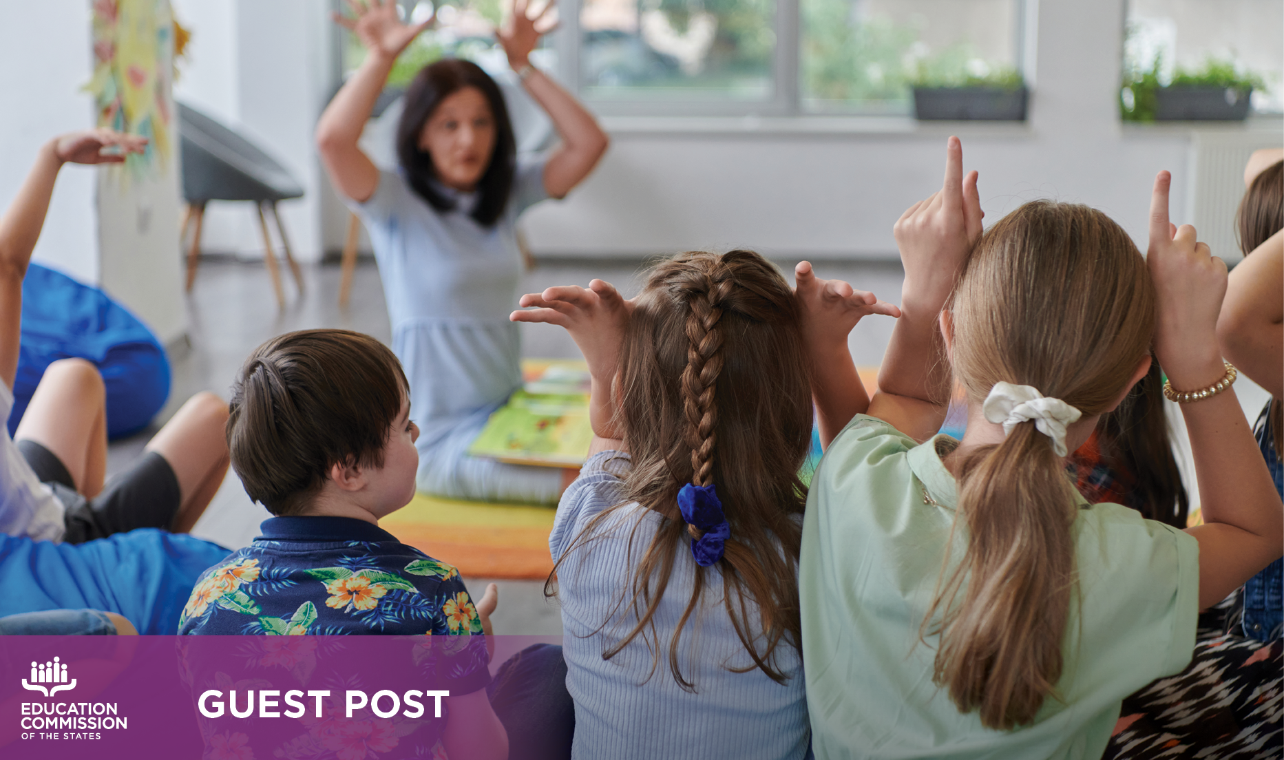 A group of young students sit in front of their teacher mimicking the teacher's movements while playing a game.