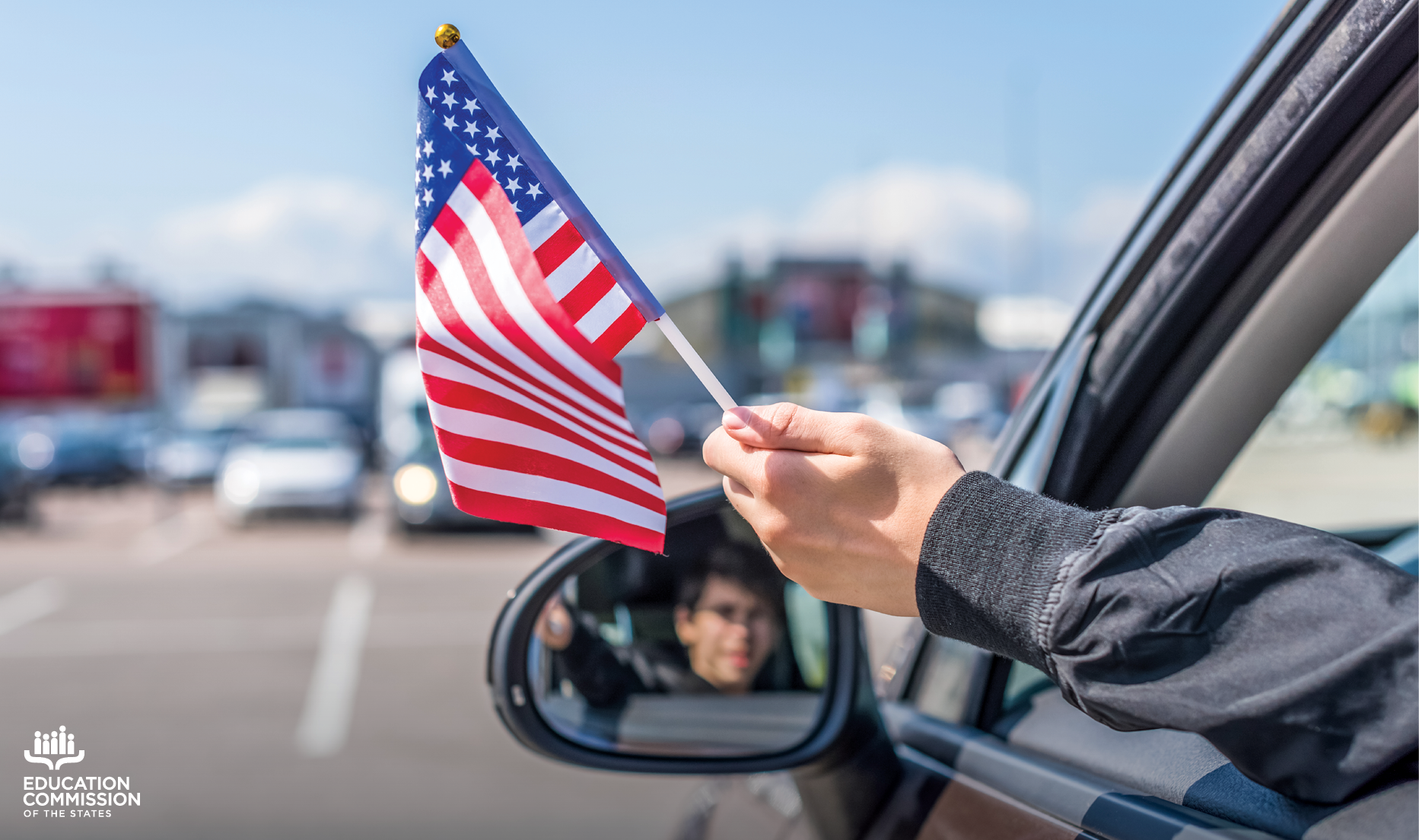 A child holds a small American flag out of a car window. The child is smiling in the reflection of the side view mirror.