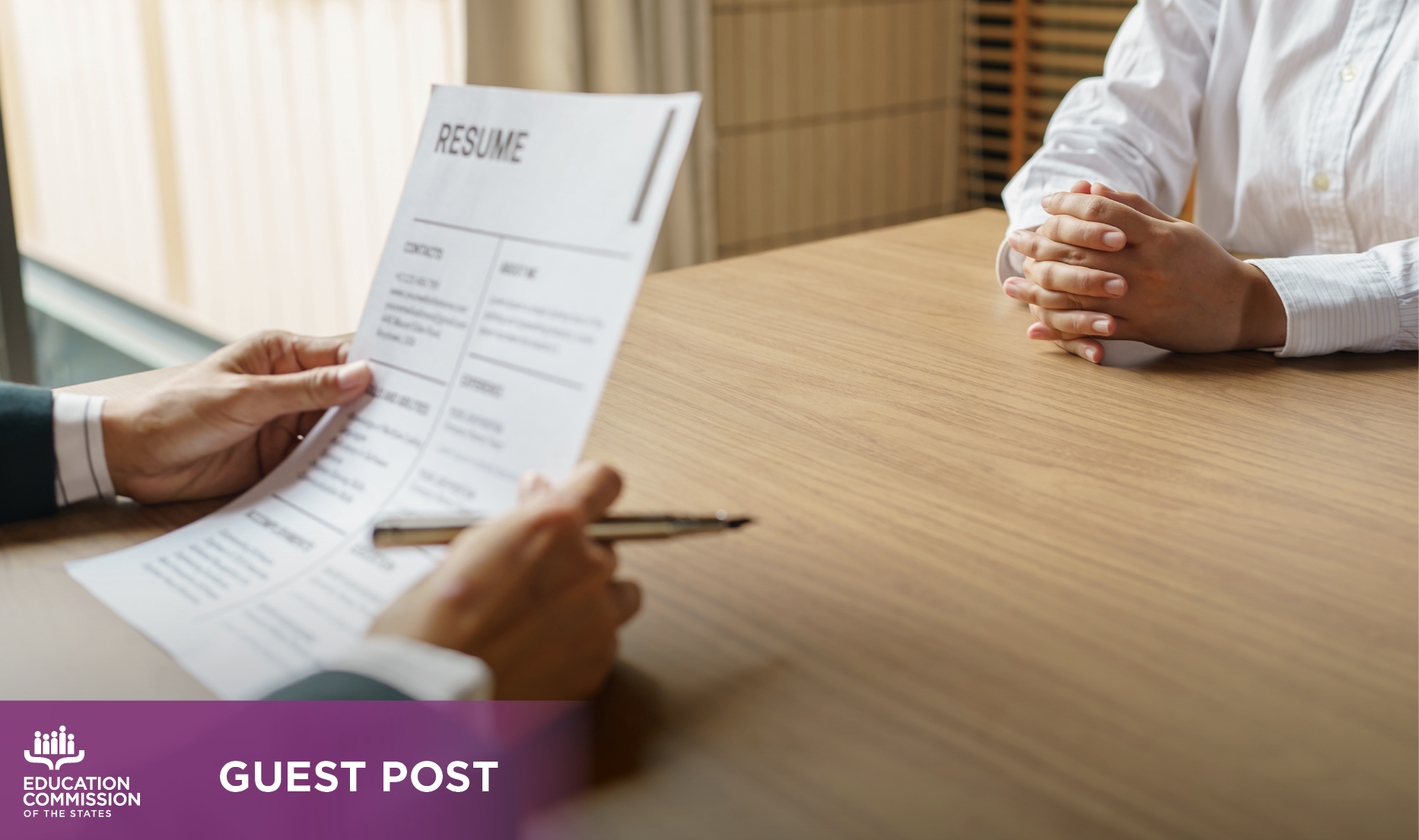 A close-up view of an interviewer and interviewee at a wooden table. The interviewer holds a resume in their hands.