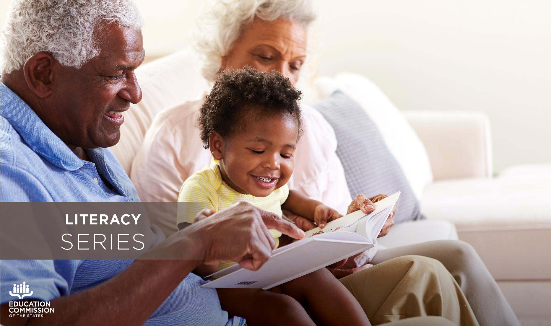 Grandparents sit on a sofa with their infant grandchild on their laps. They are all reading a storybook together.