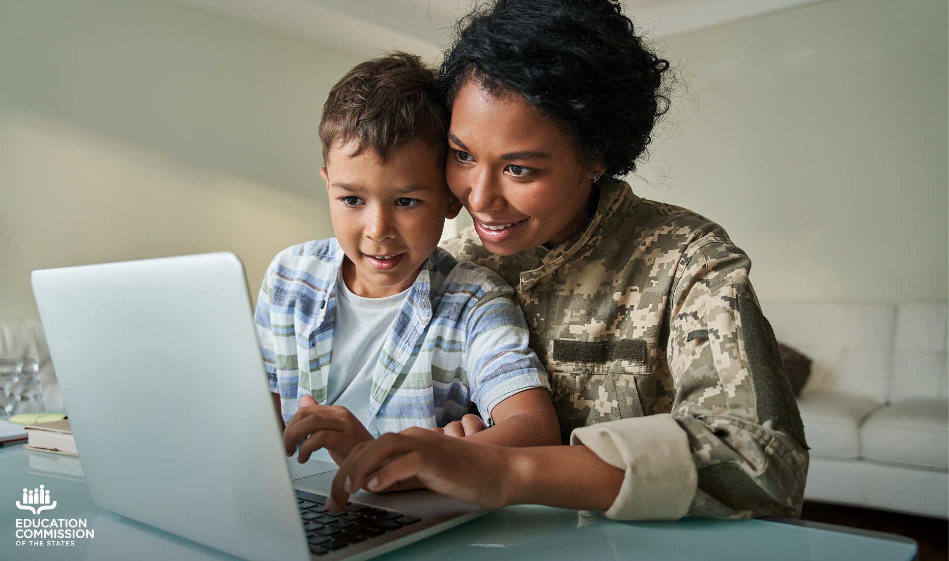 A young student sits on his mother's lap while working on a homework assignment on a laptop computer. The mother is wearing a military uniform.