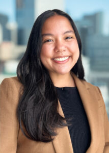 Headshot of Heena Kuwayama, smiling, wearing a camel blazer over a black shirt.