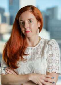A headshot of a woman with long red hair flowing down her right shoulder. She is wearing a white blouse and has a slight smile.
