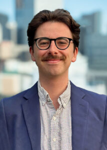 Headshot of Sam Nicholson, smiling, wearing glasses and a blue suit.