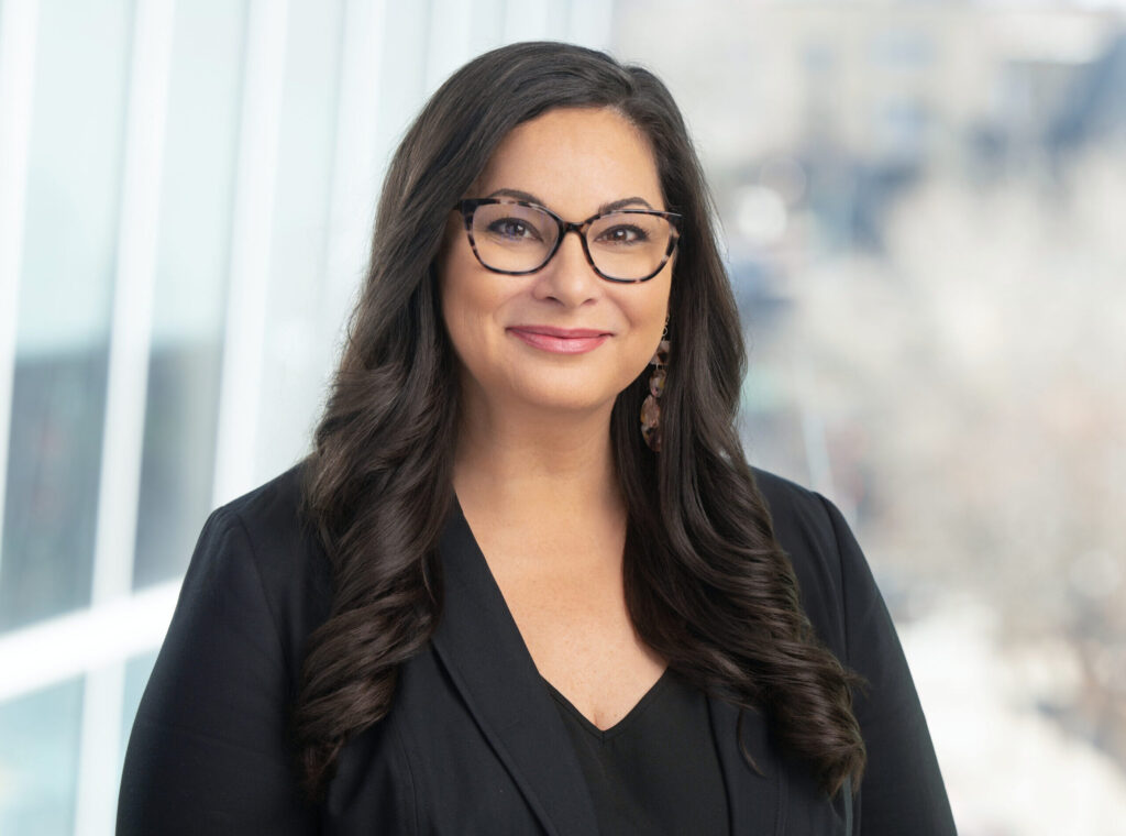 Headshot of a woman with long dark wavy hair and a subtle smile. She is wearing a black suit, dark glasses and colorful earrings.